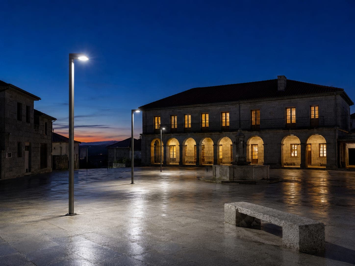 Plaza empedrada de un municipio al anochecer iluminada por farolas LED, con un edificio institucional de piedra con soportales al fondo.